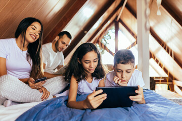 Latin family together in bed in the morning. Sister and brother using digital tablet
