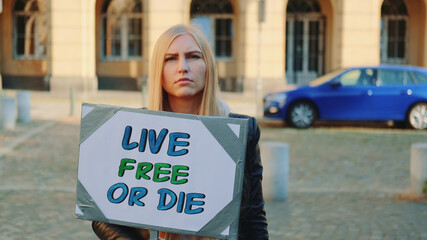 Woman with protest banner calling to live free or die. Young blonde woman walking alone in the downtown.