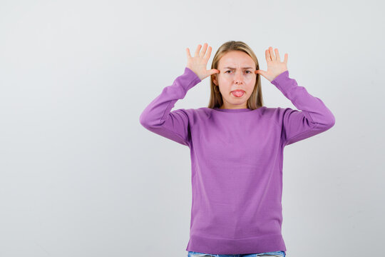 Portrait Of Young Lady Holding Hands Near Head As Ears, Sticking Tongue Out In Purple Sweater And Looking Grumpy Front View