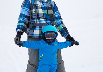 Fototapeta na wymiar Toddler boy in warm blue overalls, helmet and goggles learns to ski, holds his dad by hands. cold day in snow-capped mountains, useful activities with parents. Ski training. Sports education, care