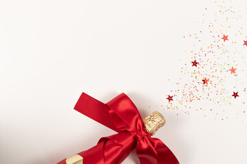 a gift bottle of champagne in golden foil wrapped in a red silk ribbon and scattered confetti on a white background