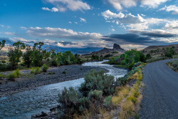 Fototapeta na wymiar Butte and river under dramatic clouds with distance mountains and trees and shrub in the foreground, Castle Rock, South Fork Shoshone River, Cody, Wyoming