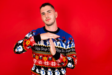 Young handsome Caucasian man wearing Christmas sweater against red wall, being upset showing a timeout gesture, needs stop, asks time for rest after hard work, demonstrates break hand sign