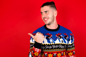 Charming Young handsome Caucasian man wearing Christmas sweater against red wall, looking at copy space having advertisements