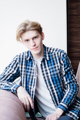 Fototapeta premium young guy with long hair, in a blue plaid shirt, casual clothes, modern hairstyle, looking at the camera, teenager sitting on devan, long hair trend, student hipster