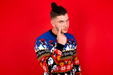 Obraz premium Young handsome Caucasian man wearing Christmas sweater against red wall, Pointing to the eye watching you gesture, suspicious expression.