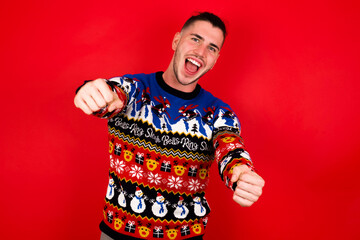 Young handsome Caucasian man wearing Christmas sweater against red wall,  imagine steering wheel helm rudder passing driving exam good mood fast speed