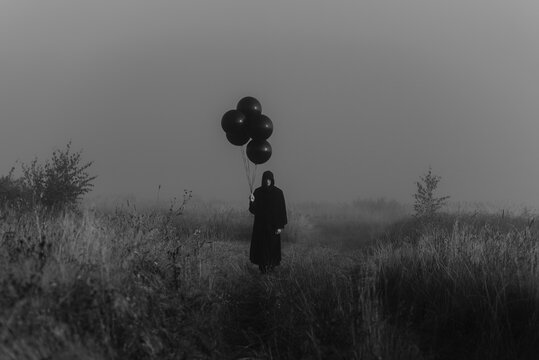 scary dark man in a monster costume with a hood with balloons in his hand