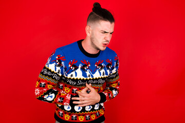 Young handsome Caucasian man wearing Christmas sweater against red wall, with hand on stomach because nausea, painful disease feeling unwell. Ache concept.