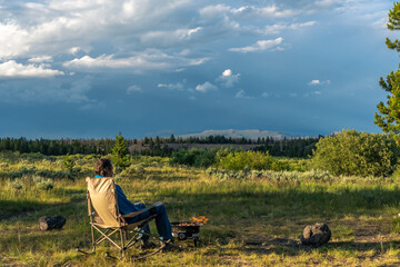 A man sitting on a rocking chair enjoying a campfire and the view of distance mountain under dramatic clouds illuminated by the setting sun, Baker's Hole Campground, West Yellowstone, Montana