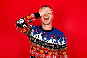 Young handsome Caucasian man wearing Christmas sweater against red wall, smiling and laughing with hand on face covering eyes for surprise. Blind concept.