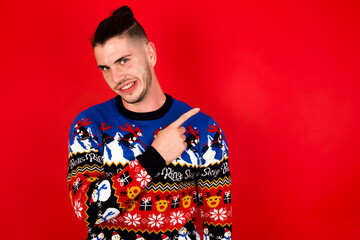 Young handsome Caucasian man wearing Christmas sweater against red wall,  Pointing aside worried...