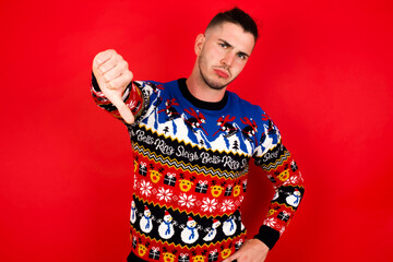 Young handsome Caucasian man wearing Christmas sweater against red wall,  feeling angry, annoyed, disappointed or displeased, showing thumbs down with a serious look