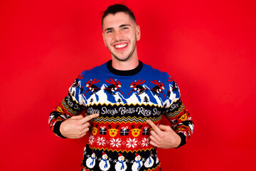 Young handsome Caucasian man wearing Christmas sweater against red wall,  points at his body, being in good mood after going shopping and making successful purchases