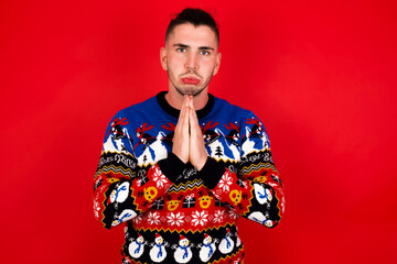 Obraz premium Positive Young handsome Caucasian man wearing Christmas sweater against red wall, smiles happily, glad to receive pleasant news from interlocutor, keeps palms together, People emotions concept.