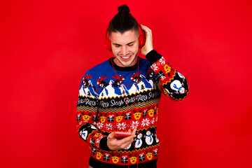 Happy Young handsome Caucasian man wearing Christmas sweater against red wall,  feels good while focused in screen of smartphone. People, technology, lifestyle