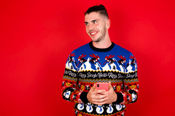 Obraz premium Young handsome Caucasian man wearing Christmas sweater against red wall, holding in hands showing new cell