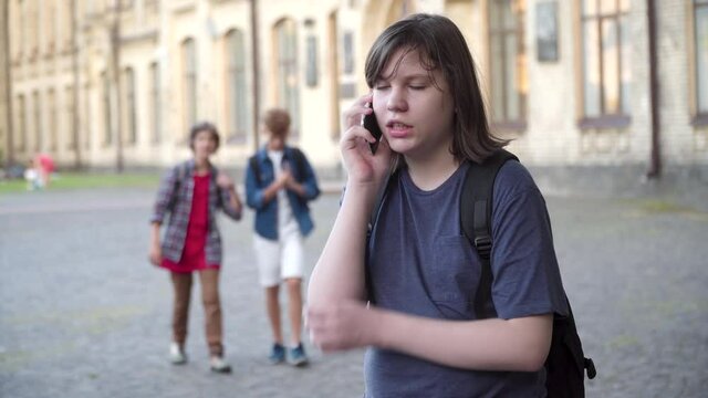 Overweight Caucasian schoolboy talking on smartphone and running away from aggressive classmates walking at the background. Portrait of scared bullied boy leaving. Bullying and fear concept.