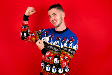 Profile photo of excited Young handsome Caucasian man wearing Christmas sweater against red wall,  isolated over yellow background raising fists celebrating black friday shopping