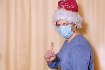 A man wearing a Santa Claus hat and a medical mask. The man makes a warning gesture. Prevention of the spread of covid-19.