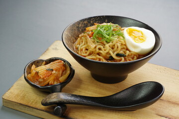Instan ramyeon or Korean noodles soup on black bowl, topped with half boiled egg, leek and Kimchi on small bowl