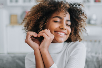 Cheerful model with perfect dark skin and white teeth hols hands in heart shape near face smiling...