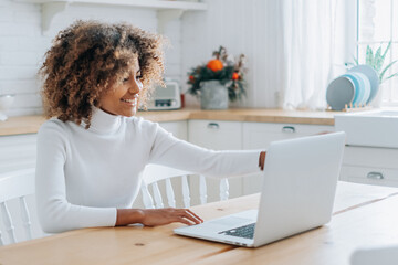 Concentrated and excited dark skinned young buyer with trendy hair looks in laptop smiles and...