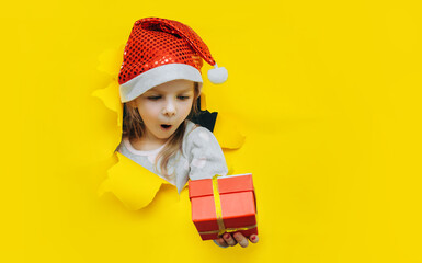 A beautiful little surprised child in a red Santa hat looks out of a hole in yellow paper and holds a box with a gift in her hand. Christmas and New Year concept. Bright background, copy space.