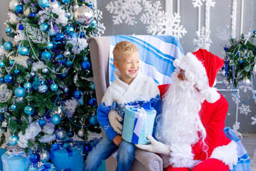 Fototapeta na wymiar Santa Claus gives a boy a gift in a blue Christmas interior near the Christmas tree, a happy child with Santa Claus