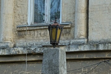 one old black iron lantern with yellow glass stands on a gray concrete pillar against the background of a house wall with a window