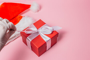 A female hand in a white medical glove opens a red gift box and pulls a white ribbon isolated on a pink background. Copy space. Flat lay. Top view