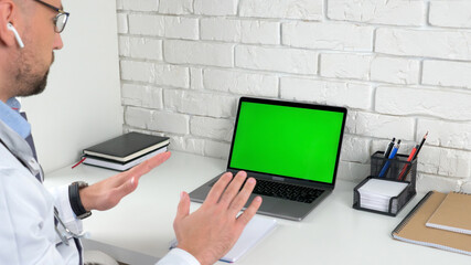 Doctor man sits on chair at desk in hospital office clinic looking green screen laptop computer talk soothes patient client with breathing technique, online remote health help, social distancing