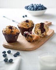 Close-up of three blueberry muffins, with one torn in half, on a wooden board on a white tablecoth in bright directional light,  Bowl of blueberries to the rear, glass of milk and napkin in foreground