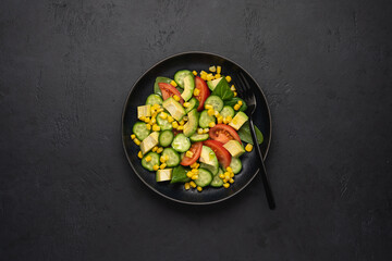 Avocado salad with corn, tomatoes, cucumbers on black slate background. Top view, copy space