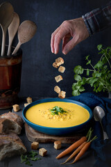 Stylised front-on shot of a bowl of carrot and coriander soup, with ingredients and bread arranged in-frame, and some croutons being sprinkled by hand above the soup bowl.