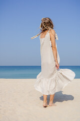 Fototapeta na wymiar A blonde smiling girl in a maxi linen dress with fluttering hair jumping and dancing on a sand beach against blue sky and sea background
