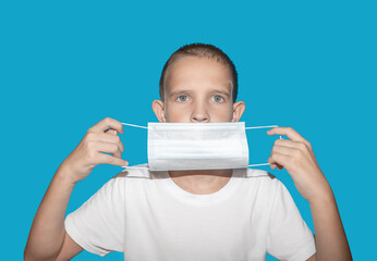 A little boy in a white T-shirt puts on a protective medical mask with two hands on a blue background.