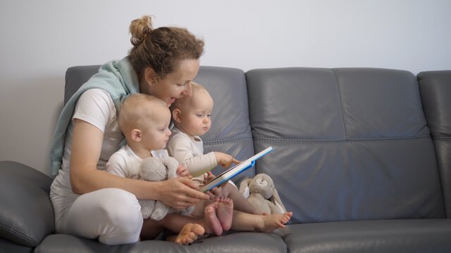 Home schooling and education for parents and children. Young mother reads book with pictures and completes exercises with two small kids on sofa