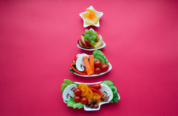 Plate in the form of a Christmas tree with vegetables, fruits, mushrooms and berries. Red background. The concept of vegetarian treats for the holiday. Copy space