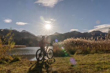 senior woman with electric mountain bike in bright backlit sun on the shore of lake Weissensee near...