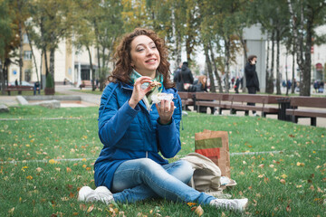 Girl student in a blue jacket eats a hamburger or cheeseburger on the street