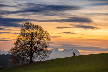 Naklejka na ściany i meble active woman riding her electric mountain bike at sunset in front of the awesome silhouette of Mount Saentis, Appenzell switzerland
