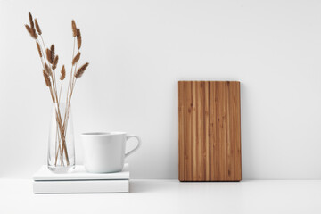 A mug on a stack of books and a transparent vase of dried flowers, a wooden board against the wall. Eco-friendly materials in interior decor. Copy space, mock up