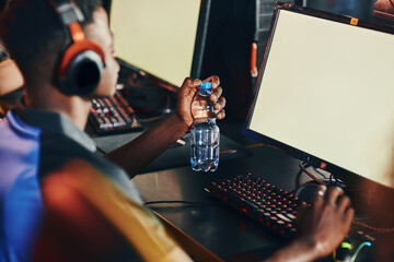 Side view of a young african guy, professional cybersport gamer wearing headphones sitting in gaming club or internet cafe and playing online video games, looking at PC screen