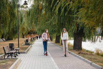 Two young beautiful girls in masks ride electric scooters in the Park on a warm autumn day. Walk in the Park.