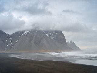 A tourst walking on the beach of Stokksnes, Mount Vestrahorn in background. Gray cloudy mood. Iceland.