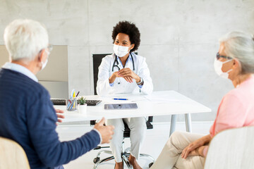 Obraz premium Senior couple with protective facial masks receive news from black female doctor