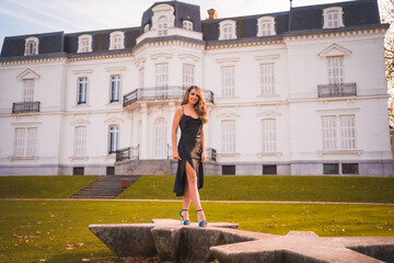 Lifestyle, blonde girl in a glamorous dress and blue high heels in a white house. Elegant outfit in autumn at sunset next to a palace in the background