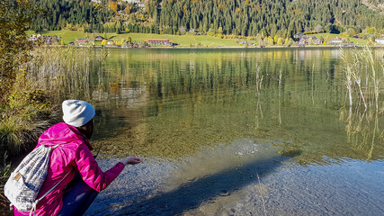 Obraz premium A woman squatting at the side of Weissensee and playing with the cold water. She wears a pink jacket and a beanie. The surface of the lake is calm and reflecting the Alps. Joyfulness and happiness.