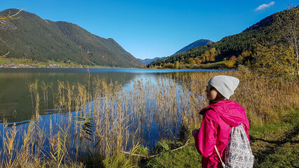 A woman walking along the Weissensee lake in Austria. She wears a pink jacket and a beanie. The surface of the lake is calm and reflecting the Alps, high grass at the shore. Joyfulness and happiness.